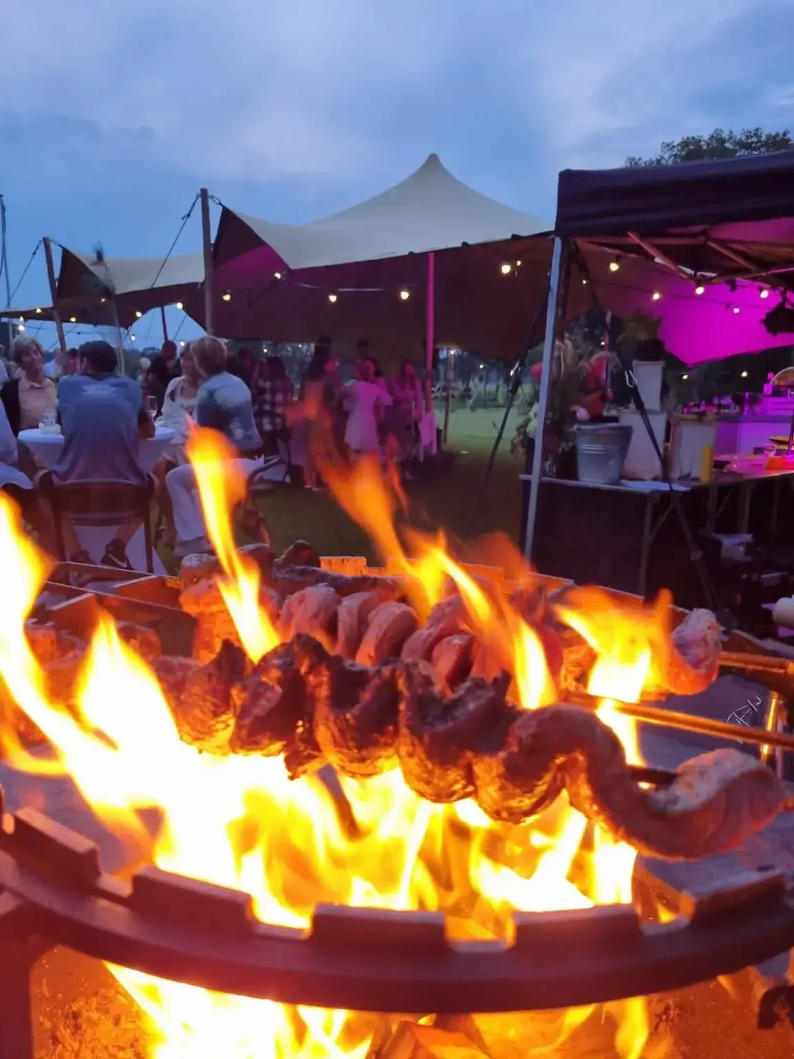 Vlammen slaan op uit een Ofyr met spiesen, midden tussen de gasten onder een partytent in de schemering